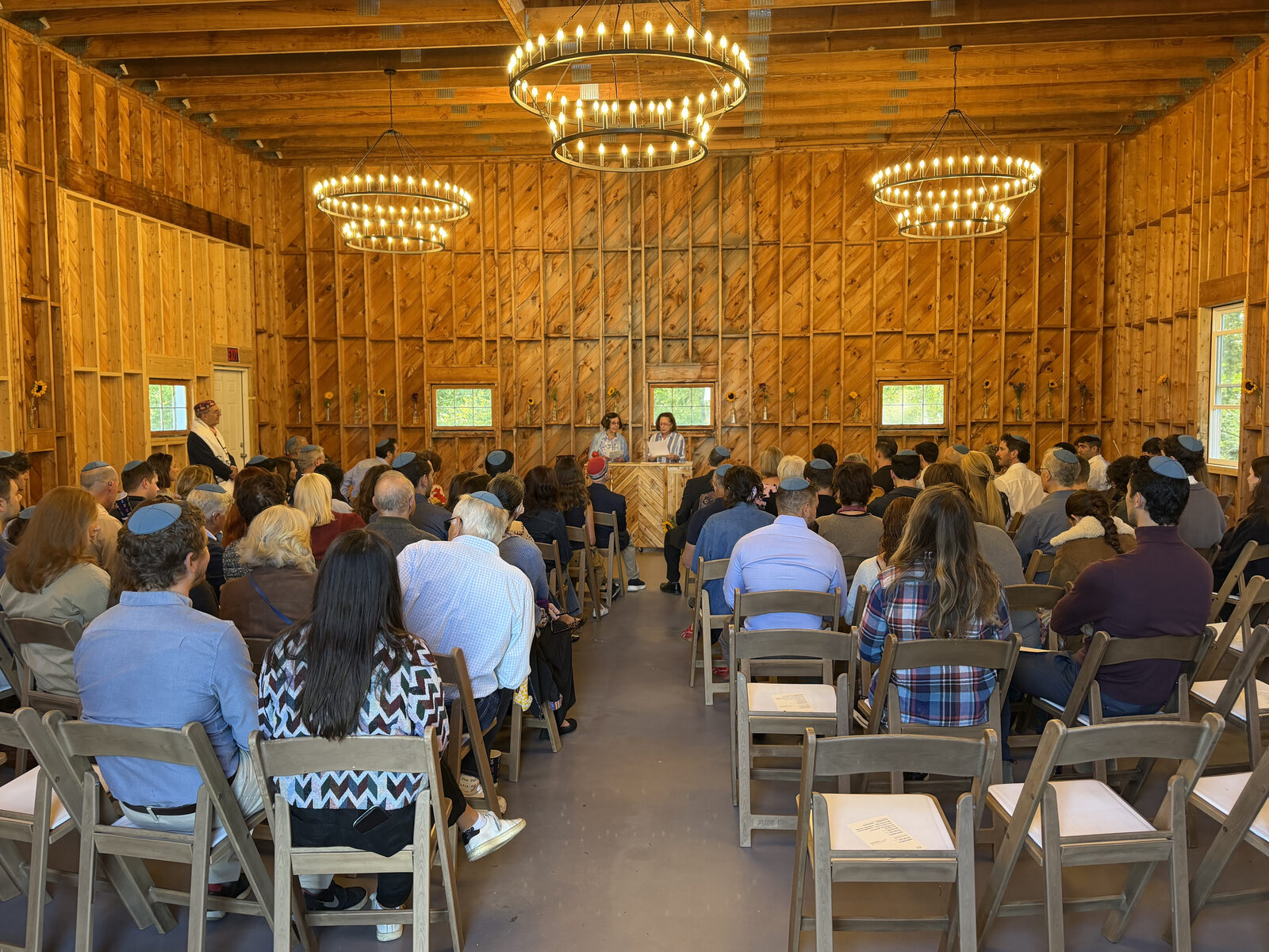 Guests seated in rows inside the barn for a ceremony, wagon-wheel chandeliers glowing overhead
