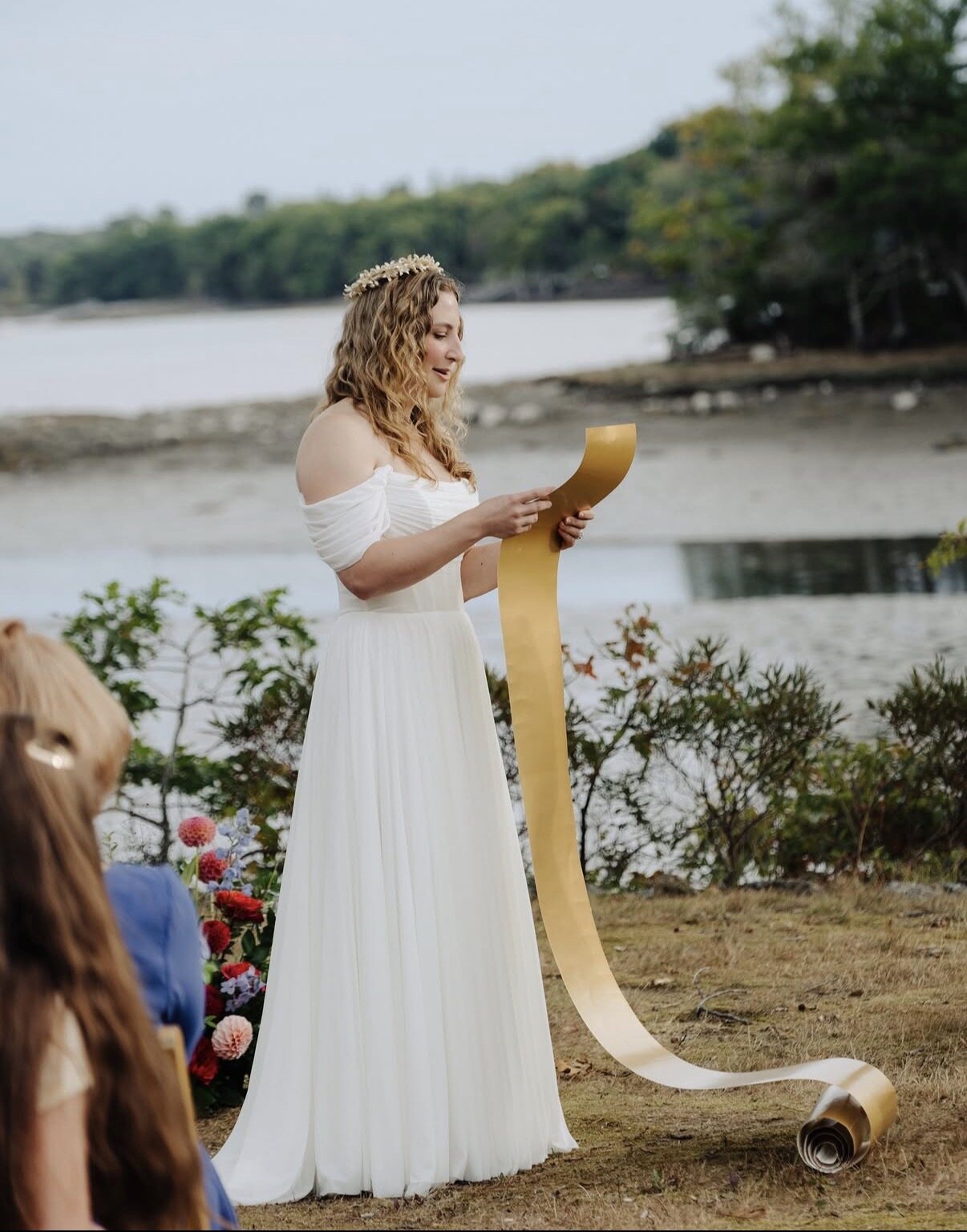 A bride reads her vows from a long gold scroll at the water's edge, the harbor behind her