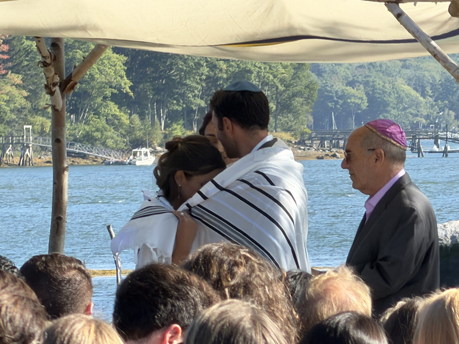 A couple wrapped in a tallit stands beneath a chuppah at the water's edge as guests look on