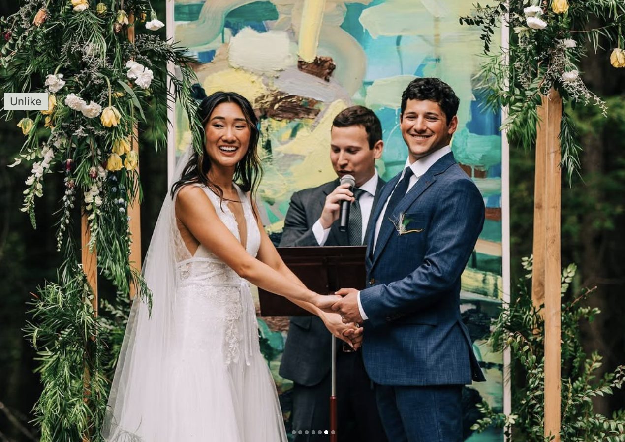 A couple holds hands at the altar in front of a painted backdrop, flanked by floral arches