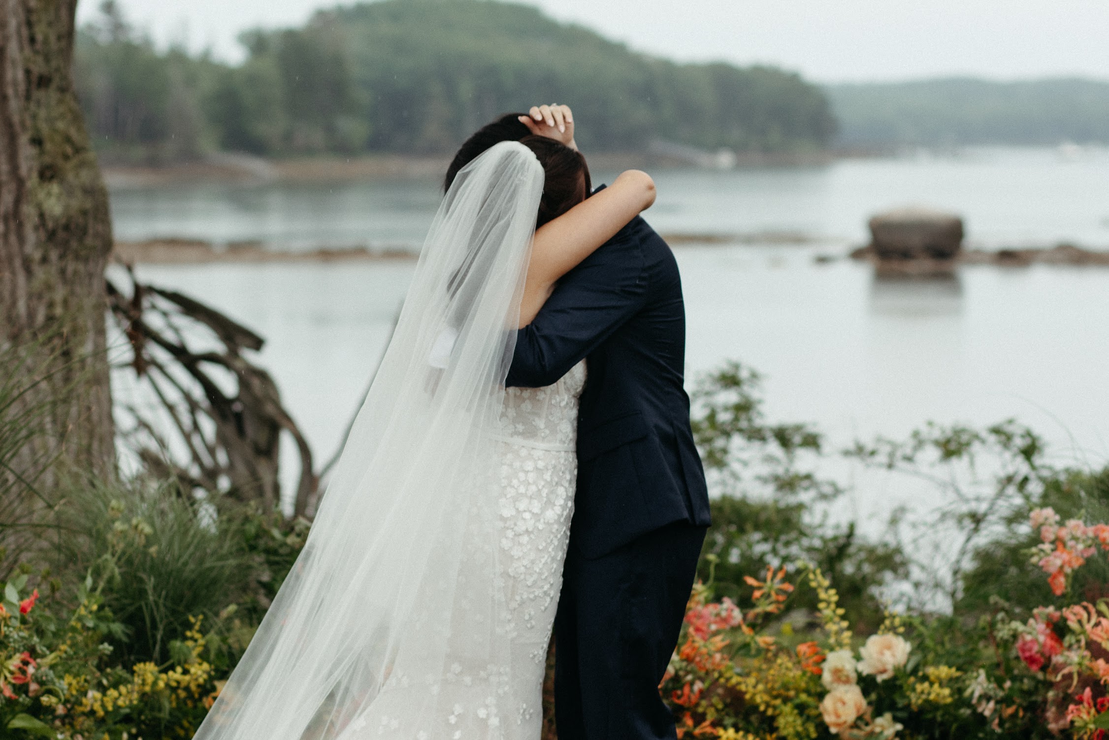A bride and groom embrace under a veil on a misty, rainy day by the water