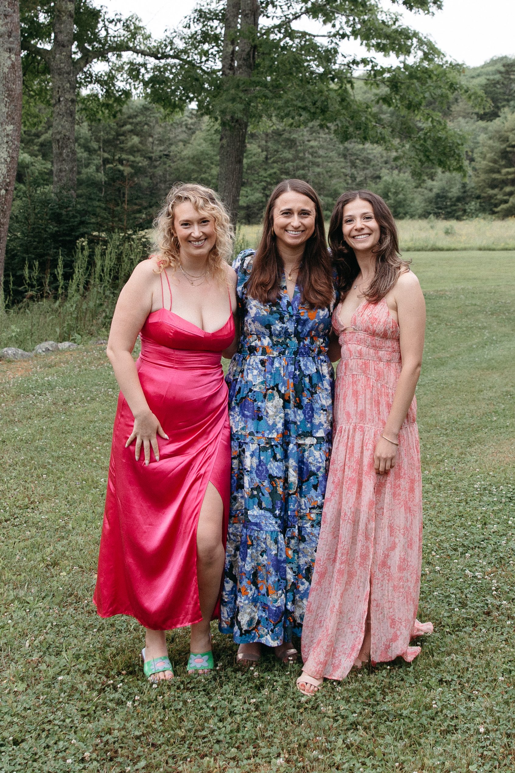 Three bridesmaids in colorful gowns smiling together at the edge of the field