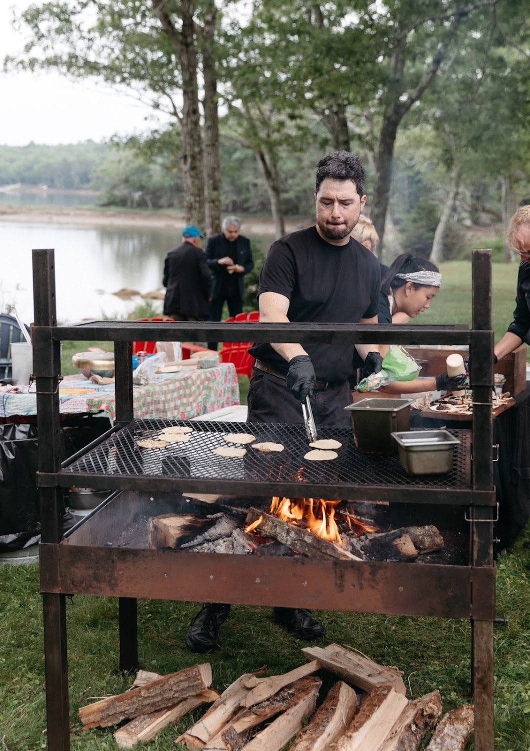 A chef grills over an open fire at the water's edge during cocktail hour