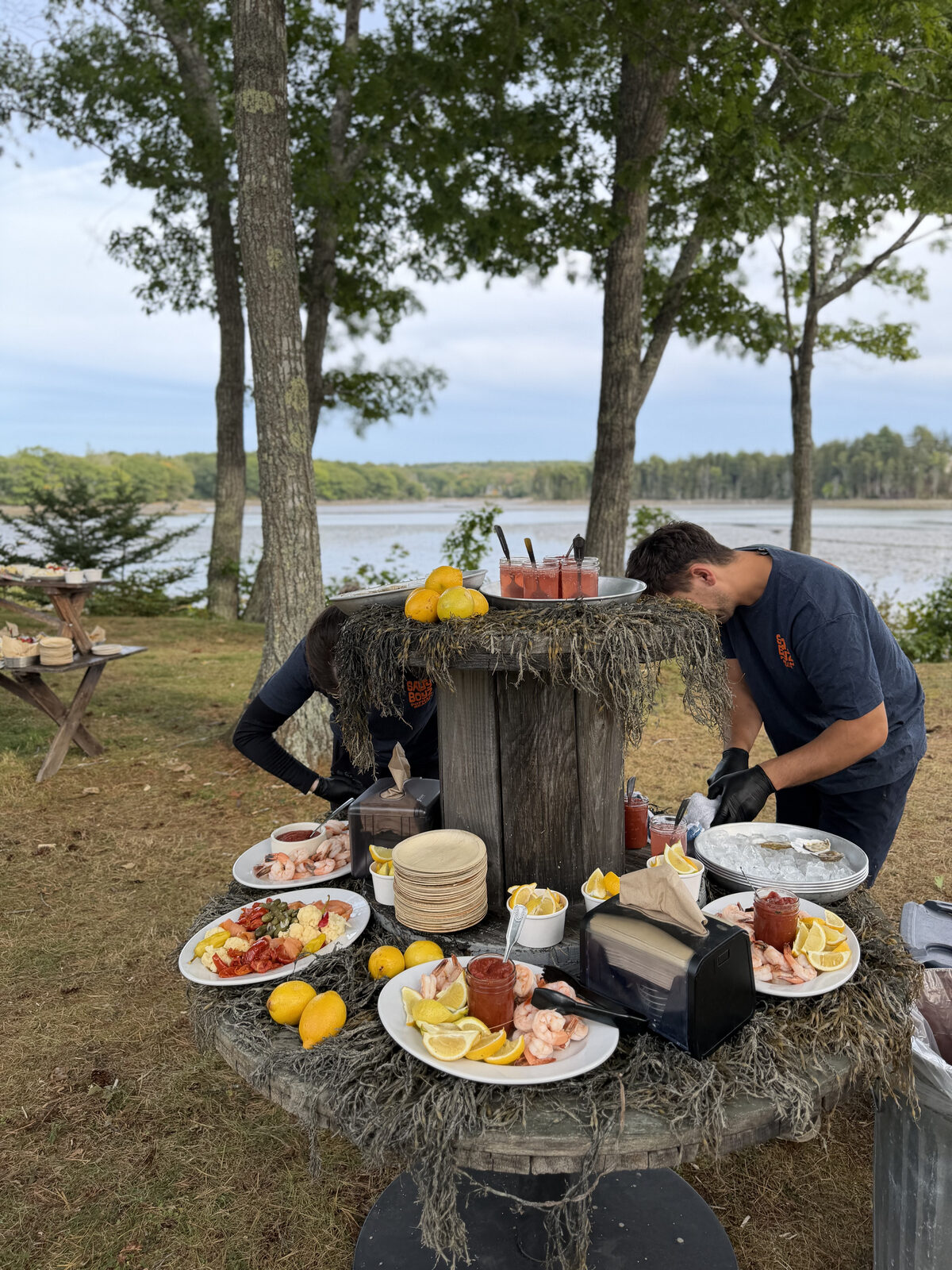 Caterers set out a raw bar of shrimp, oysters, and lemons on a seaweed-draped spool table beside the harbor