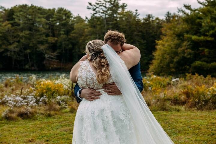 A couple embraces on the lawn near the water, surrounded by wildflowers