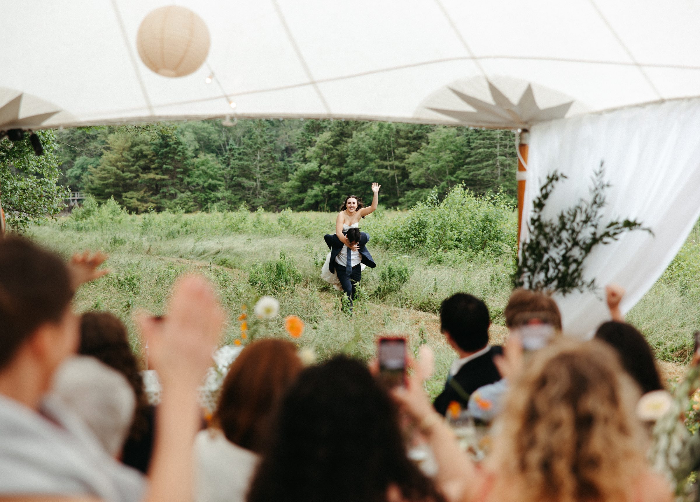 A newlywed couple runs through a mown path in the meadow toward the reception tent, guests cheering