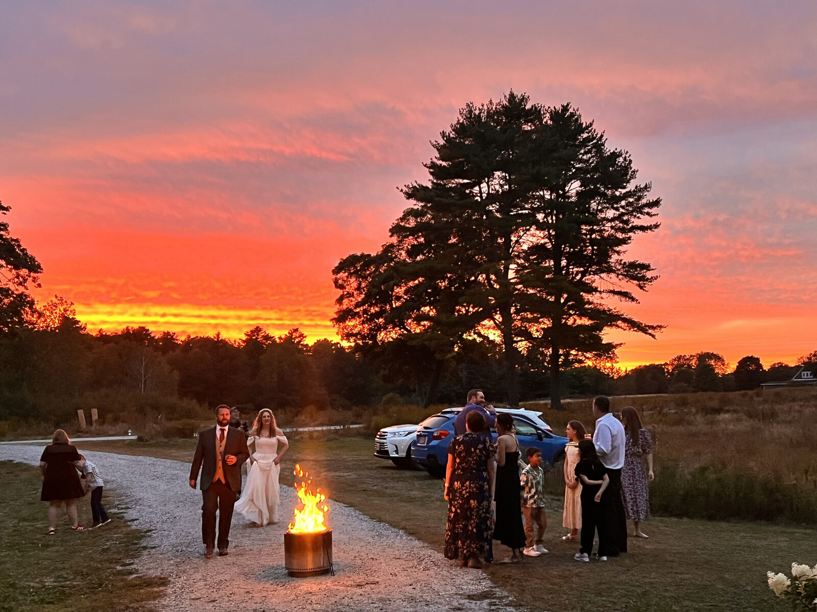A bride and groom walk past a fire pit as guests gather on the drive under a blazing orange sunset