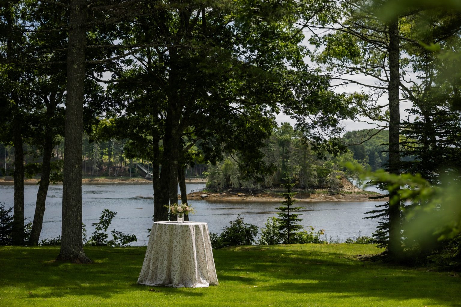 A cocktail table dressed in floral linen set beneath shade trees along the water