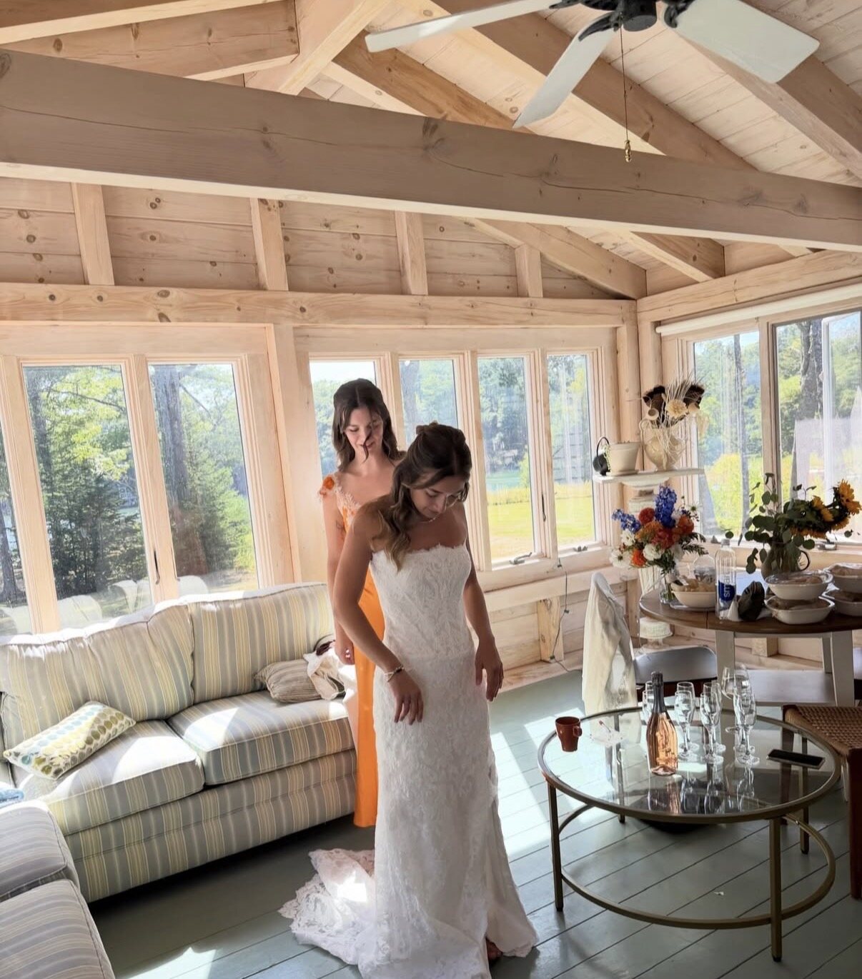 A bridesmaid laces up the bride's lace gown in the sun-filled sunroom, flowers and champagne glasses on the table beside them