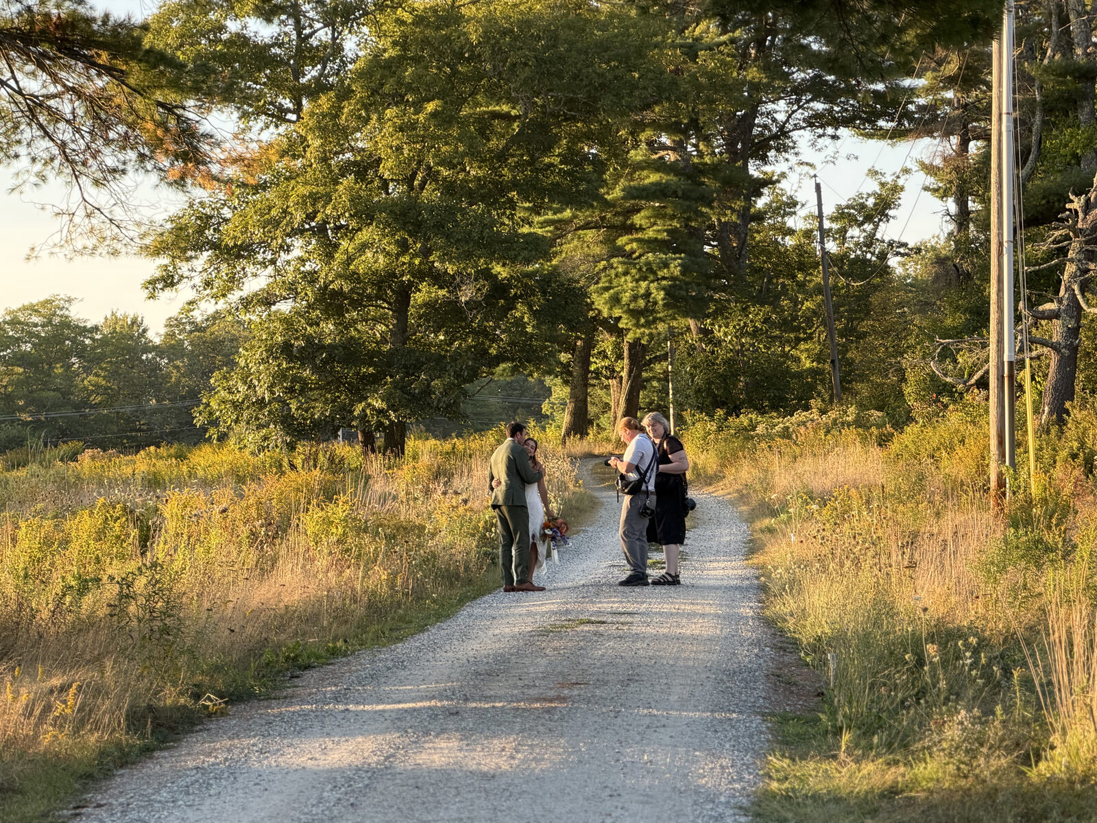 A couple walks the gravel path through golden meadow grass and tall trees in late-day light