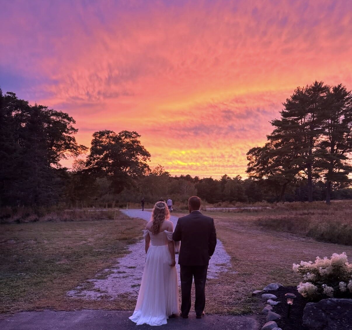 A bride and groom stand at the head of the gravel path, looking out at a blazing pink and orange sunset over the meadow